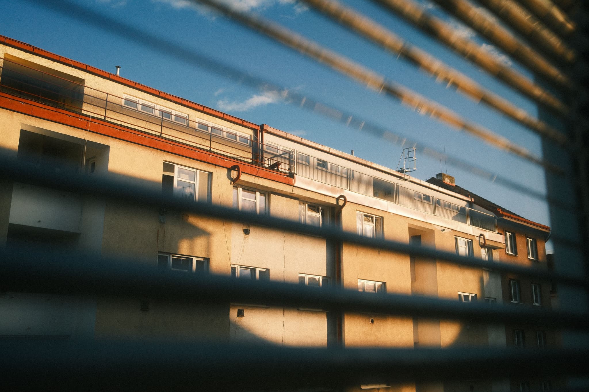 A view of a building with a red roof and multiple windows, seen through partially open blinds, under a blue sky
