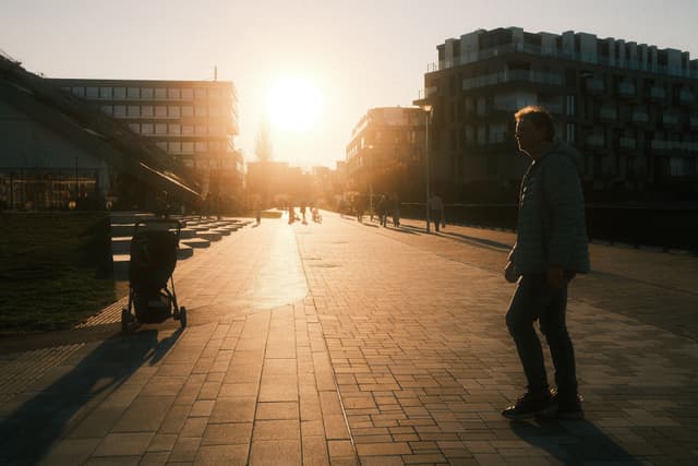 A person walking on a sunlit pathway with buildings on either side, and a stroller in the foreground