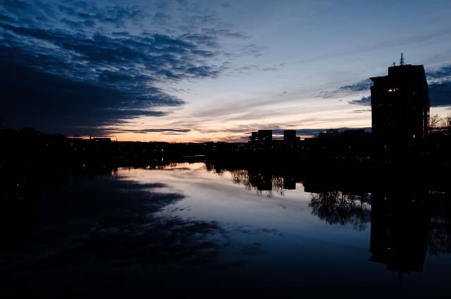 A serene river scene at dusk with a silhouetted city skyline and dramatic clouds reflecting on the water