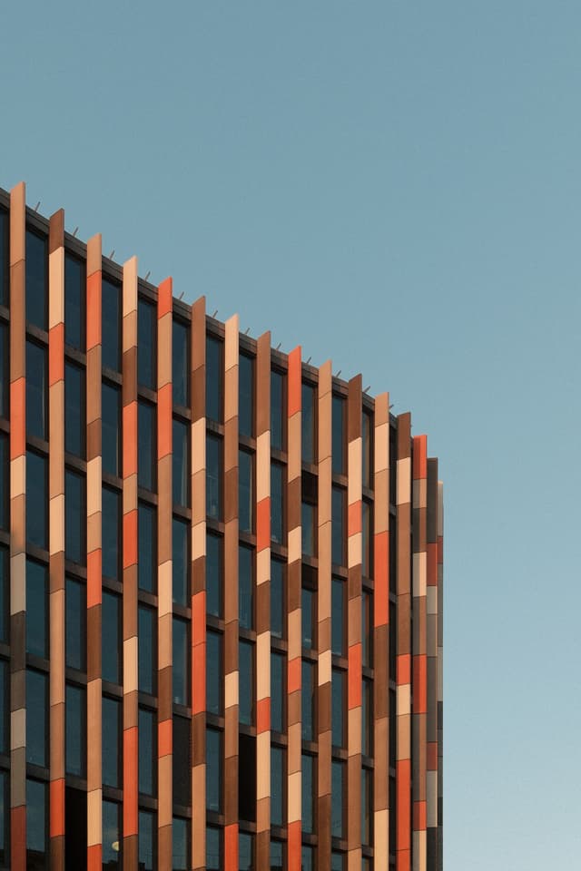 A modern building facade with a pattern of vertical panels in shades of red, orange, and beige against a clear blue sky