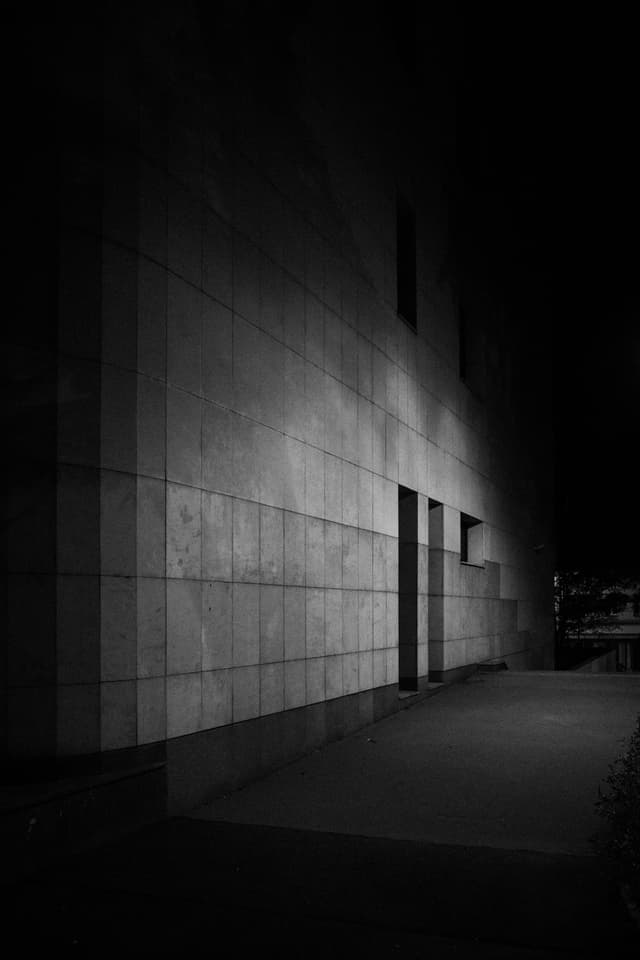 A dimly lit, monochrome scene of a building's exterior wall with geometric patterns, featuring a sidewalk and a few windows