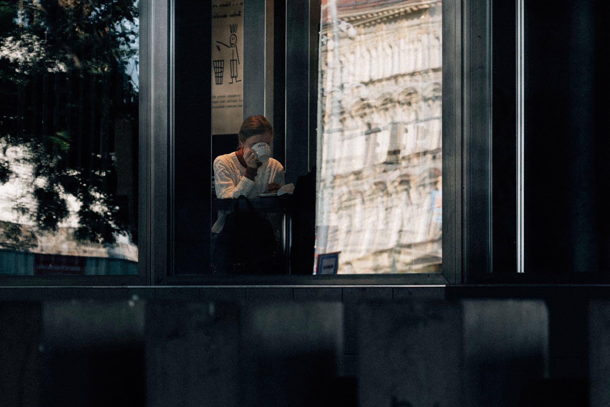 A person sitting inside a building, viewed through a window, with reflections of an exterior structure visible on the glass