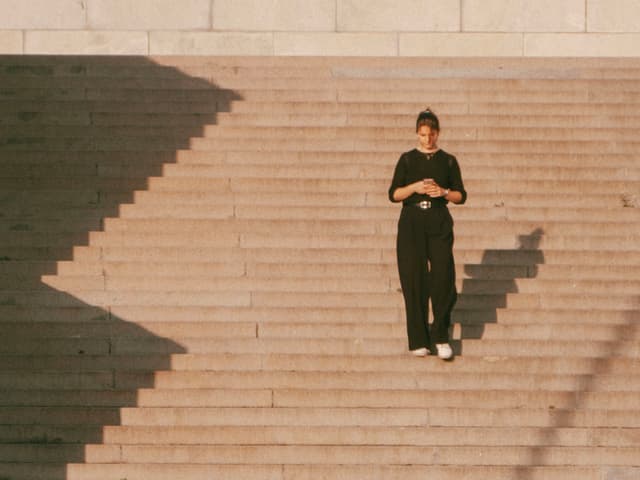 A person in dark clothing stands on wide stone steps, with long shadows cast by the sunlight