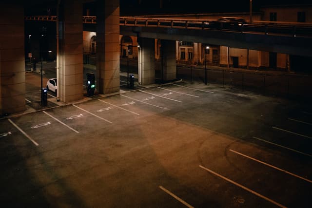 An empty parking lot at night, illuminated by streetlights, with an overpass and a few parked cars in the background