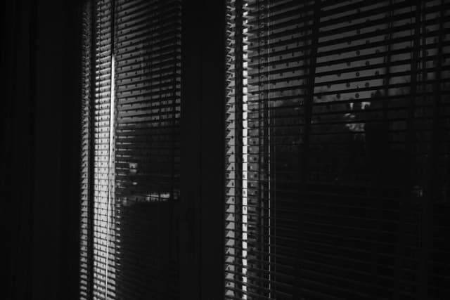 A dimly lit room with vertical blinds partially open, allowing light to filter through, creating patterns of light and shadow