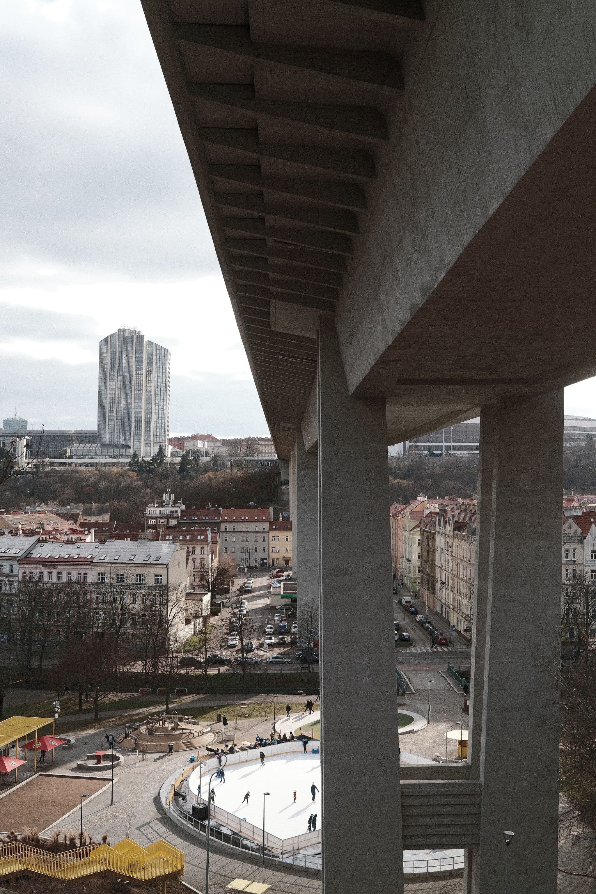 A large concrete bridge spans over a cityscape with a tall building in the background and a circular plaza below