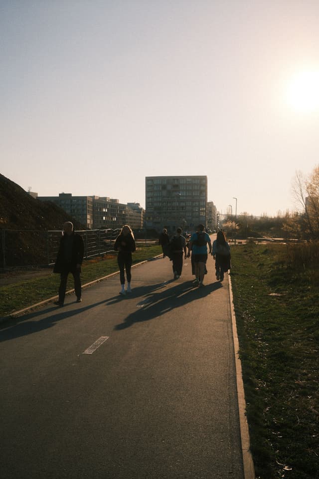 A group of people walking on a paved path with long shadows cast by the setting sun, surrounded by grassy areas and buildings in the background