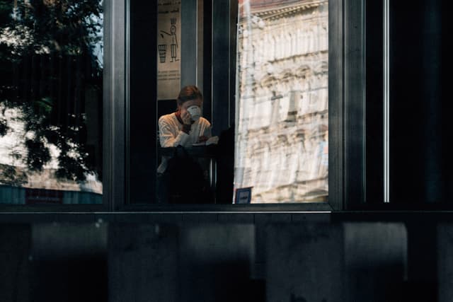A person sitting inside a building, viewed through a window, with reflections of an exterior structure visible on the glass