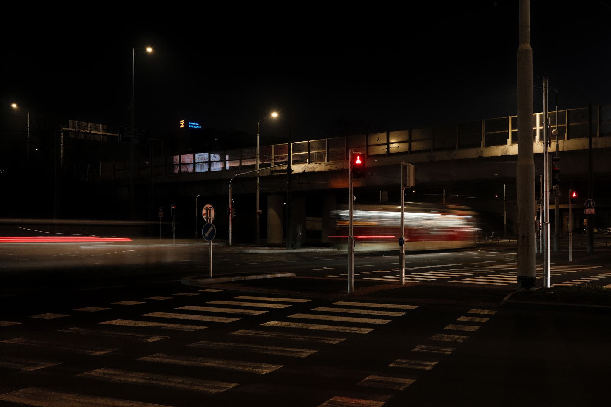 A nighttime urban scene with a blurred vehicle passing under a bridge, illuminated by streetlights and a red traffic signal