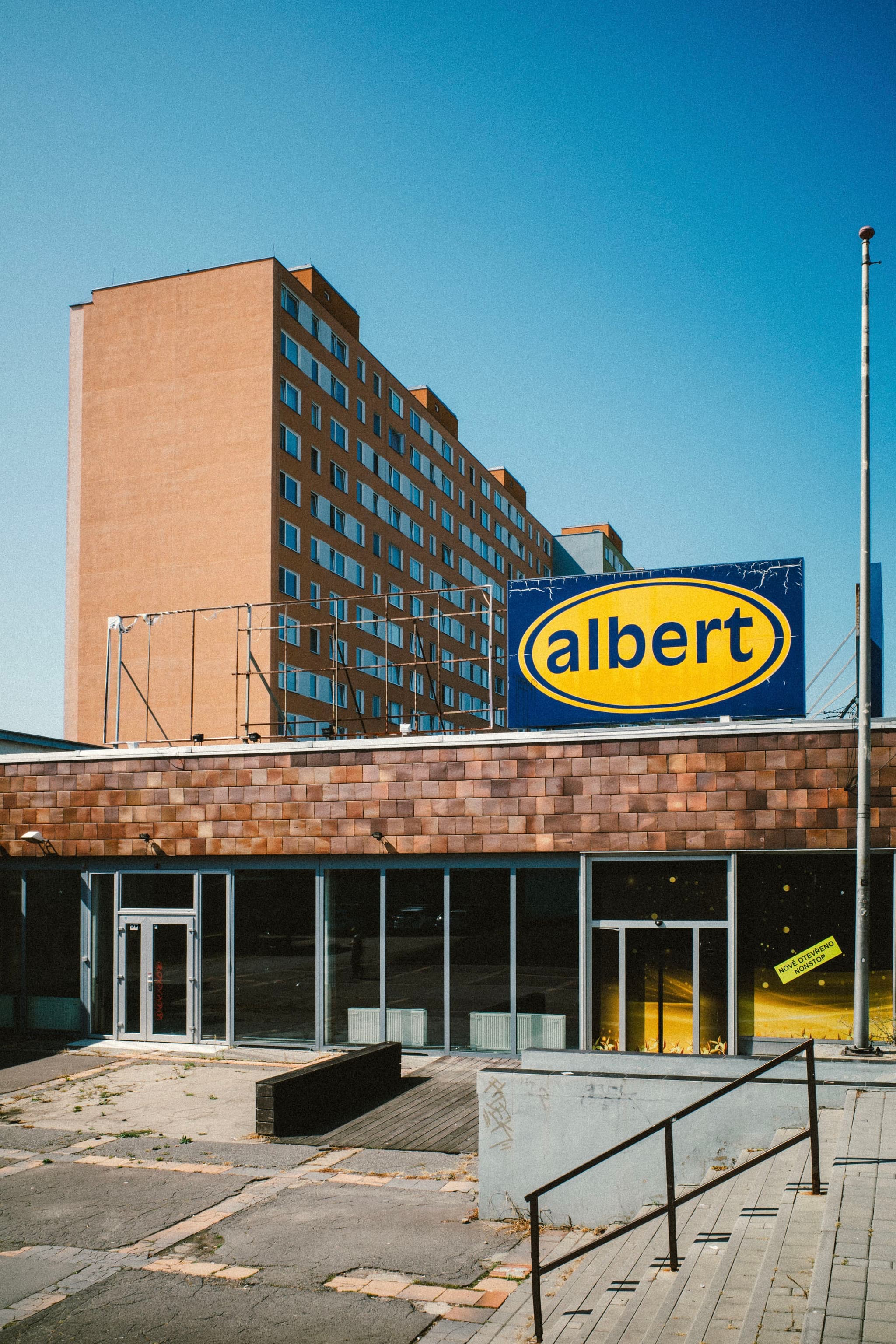 A building with a large albert sign in front, featuring a brick facade and a multi-story apartment block in the background under a clear blue sky