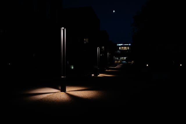 A dimly lit pathway at night with streetlights casting pools of light on the ground, surrounded by dark buildings and a small crescent moon in the sky