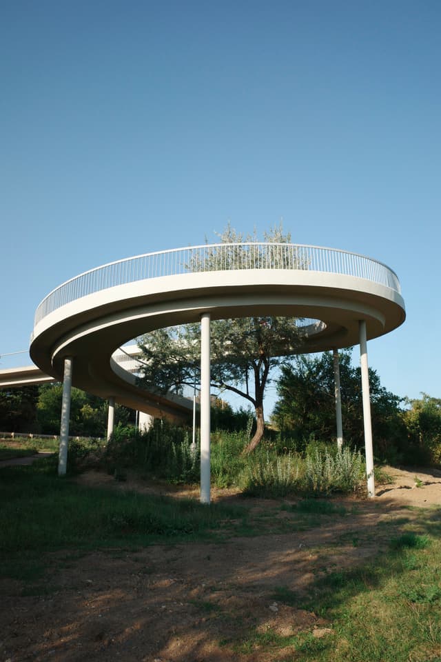 A circular elevated walkway with metal railings, supported by pillars, surrounded by greenery and trees under a clear blue sky