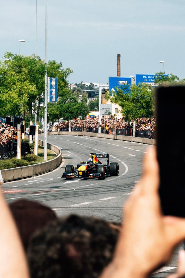 A Formula 1 car is racing on a city street, surrounded by a large crowd of spectators Trees and buildings are visible in the background, and people are capturing the moment with their phones