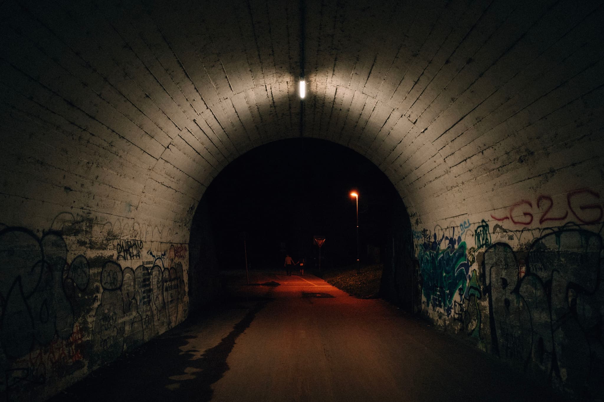 A dimly lit tunnel with graffiti on the walls, leading to a dark path illuminated by a distant streetlight