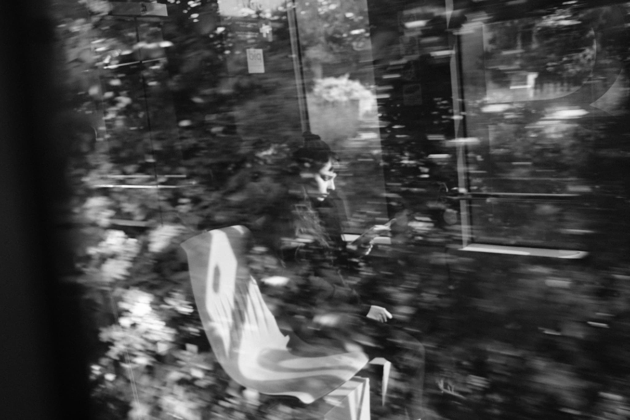 A black and white scene with a person sitting on a bench, surrounded by blurred reflections and foliage, creating an abstract, dreamlike effect
