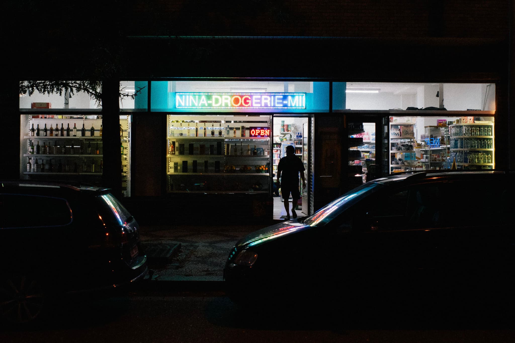 A dimly lit street scene with a person entering a brightly illuminated store, flanked by parked cars