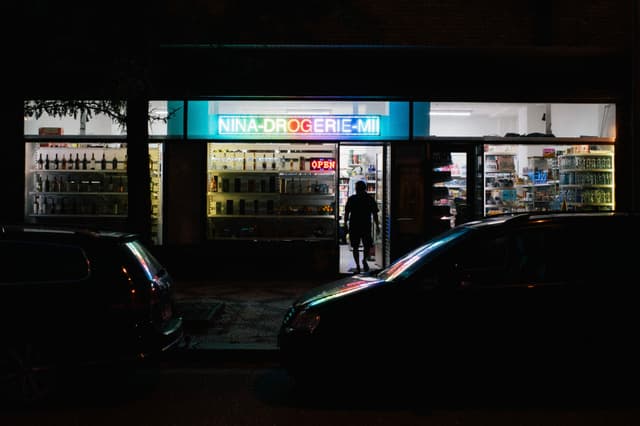 A dimly lit street scene with a person entering a brightly illuminated store, flanked by parked cars