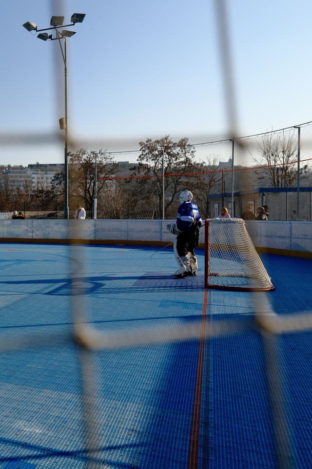 A hockey goalie stands in front of a net on an outdoor rink, viewed through a protective netting. The rink has a blue surface, and there are trees and buildings in the background under a clear sky