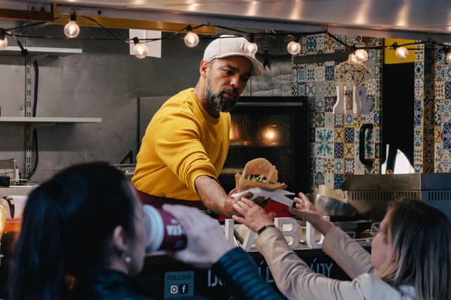 A man in a yellow sweater and cap serves food from a food truck to two women