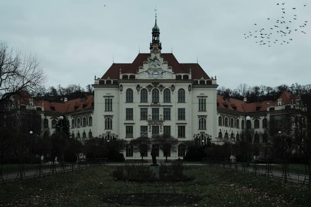 A large, ornate building with a red roof and multiple windows, surrounded by trees and a garden, under a cloudy sky with birds flying