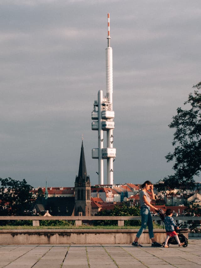 A tall, modern tower stands prominently against a cloudy sky, with a church spire and rooftops in the foreground A person walks with a child in a stroller on a paved area, and a tree is visible on the right