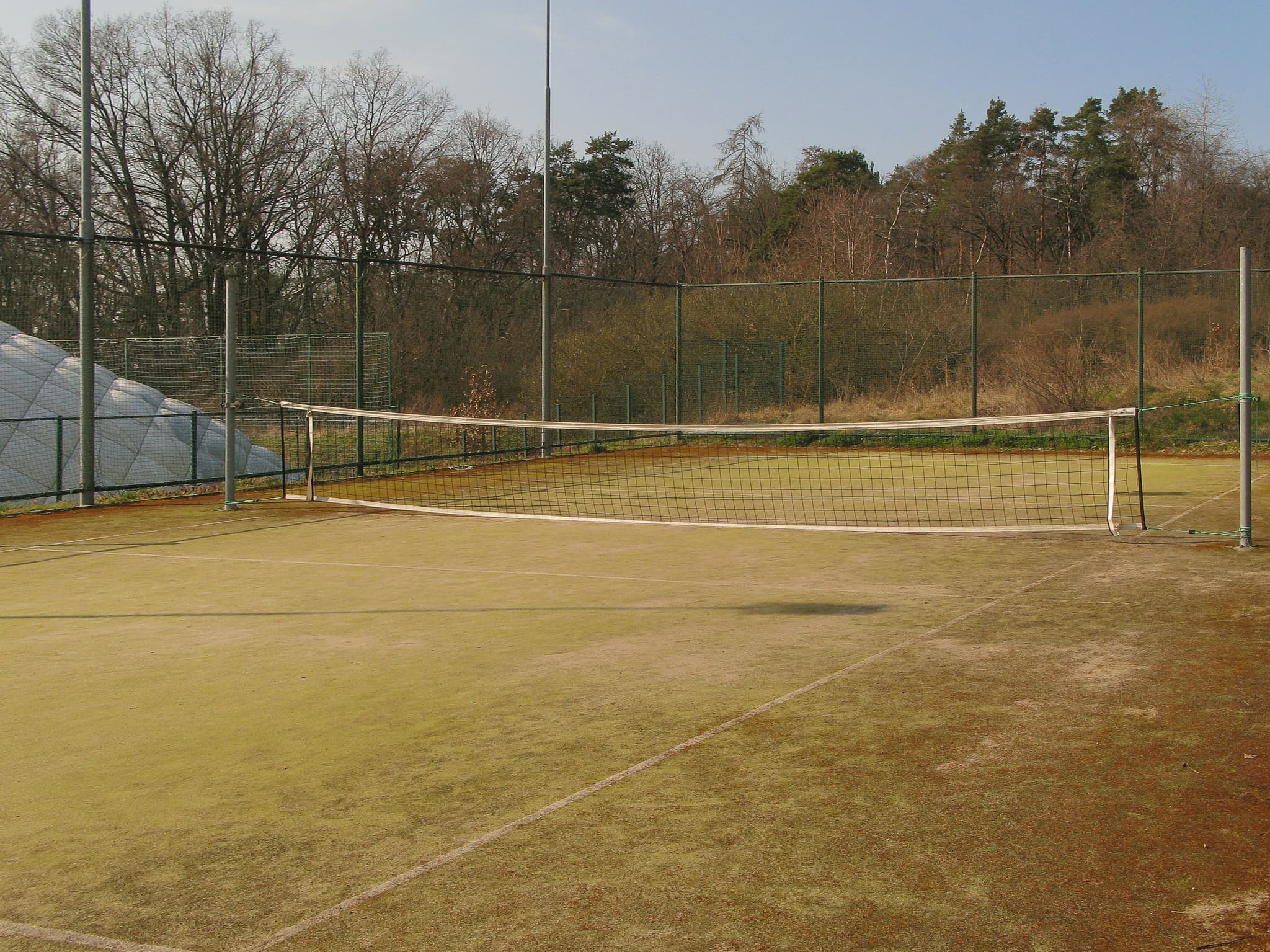 An outdoor tennis court with a net, surrounded by a fence and trees in the background The court surface appears worn and discolored