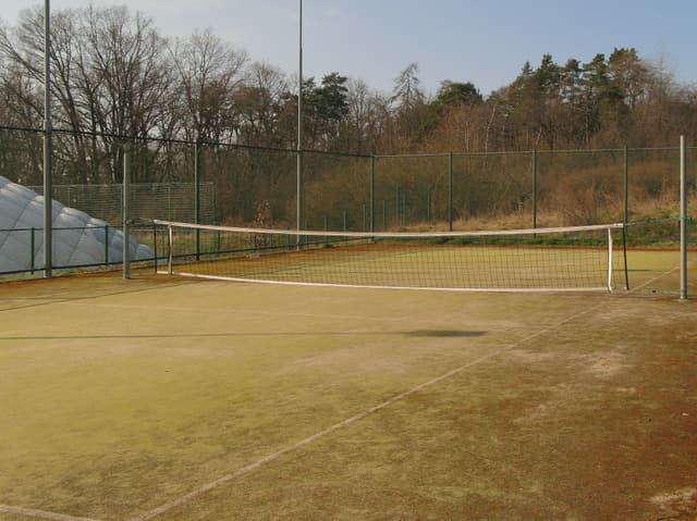 An outdoor tennis court with a net, surrounded by a fence and trees in the background The court surface appears worn and discolored