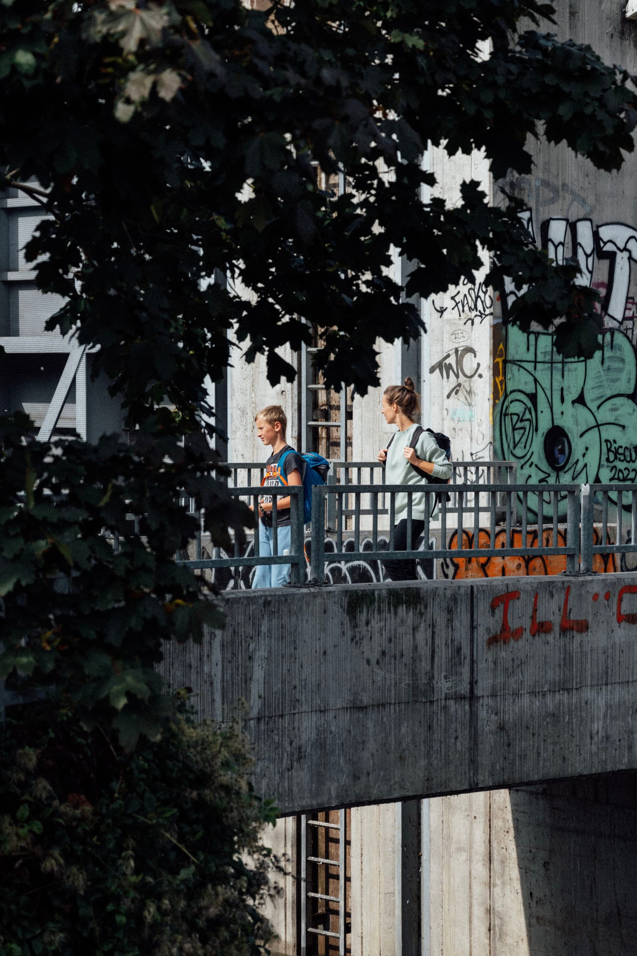 Two people walking on a graffiti-covered bridge, partially obscured by tree leaves