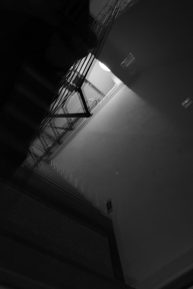 A dimly lit, black and white staircase with a railing, viewed from an angle, creating a dramatic and abstract effect