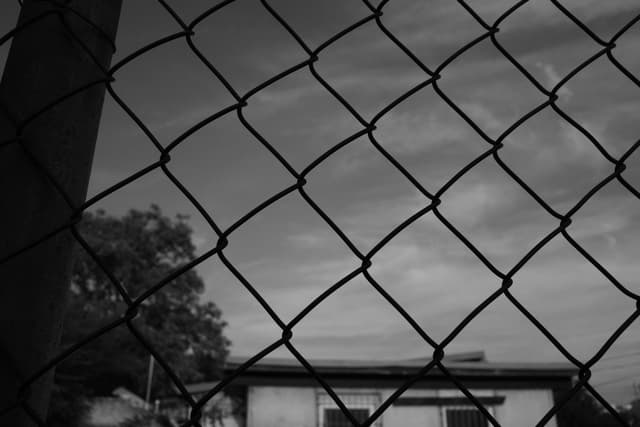 A black and white photo of a chain-link fence with a building and tree in the background