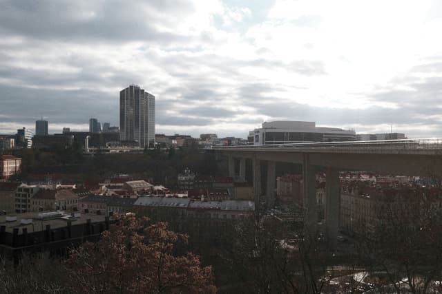 A cityscape featuring a tall building, a bridge, and several smaller structures under a cloudy sky