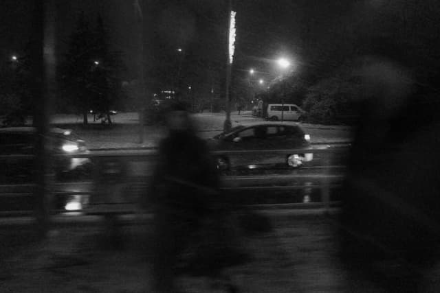 Blurry silhouettes of pedestrians walking along a wet street at night with cars and streetlights in the background