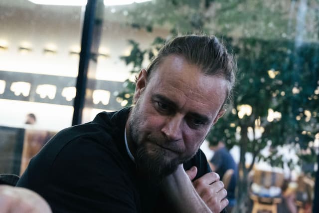 A man with a beard and a bun, wearing a black shirt, is sitting indoors with trees and people in the background