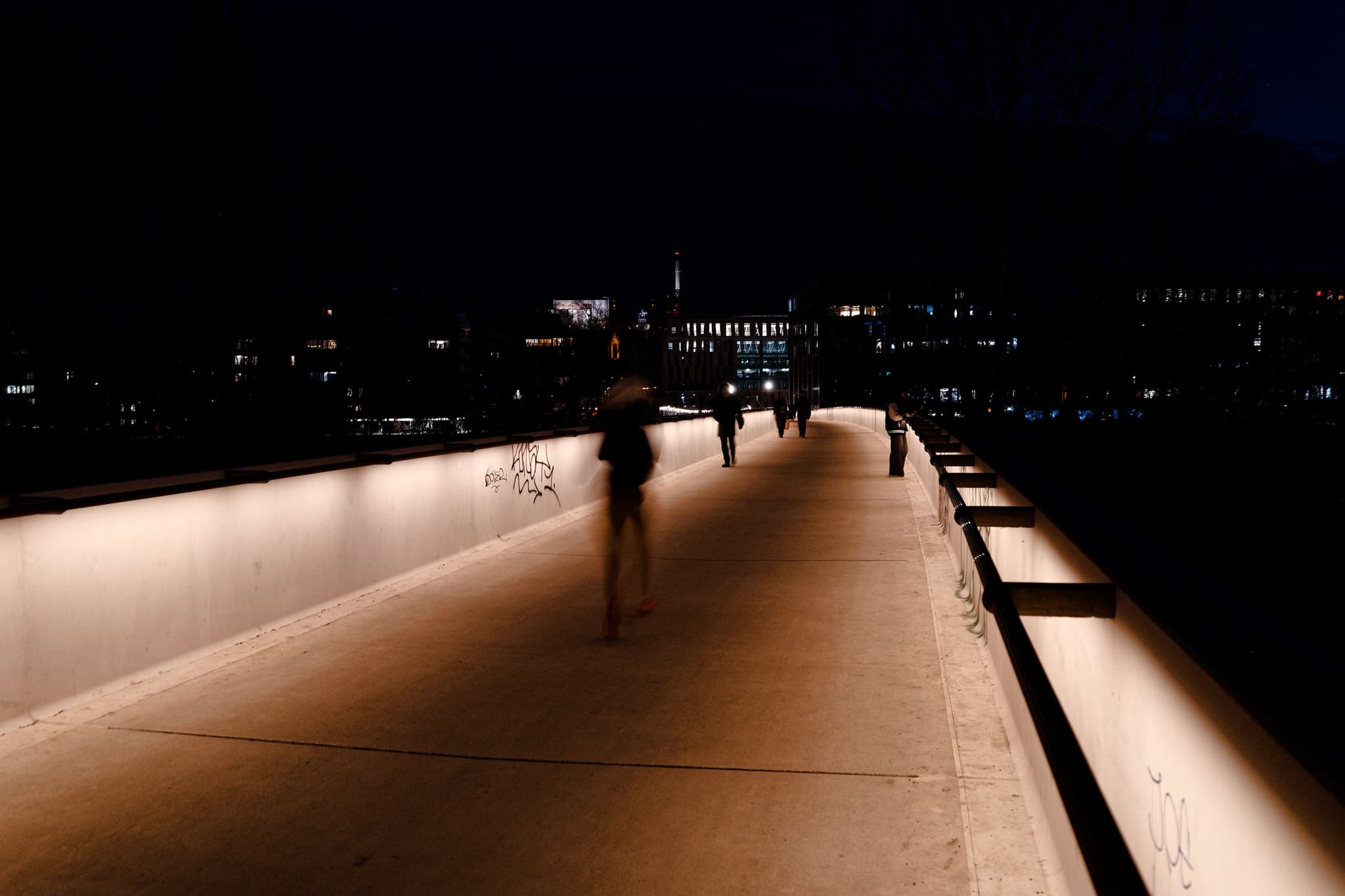 A dimly lit bridge at night with blurred figures walking along it, surrounded by city lights in the background