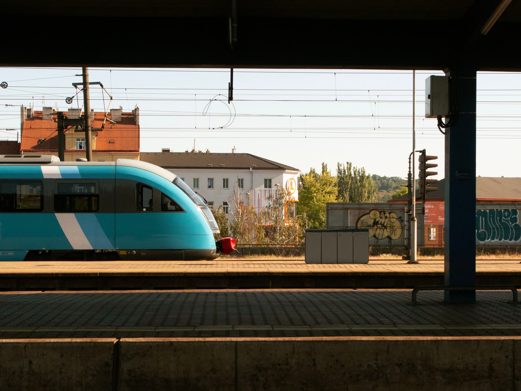 A blue train at a station platform with buildings and power lines in the background