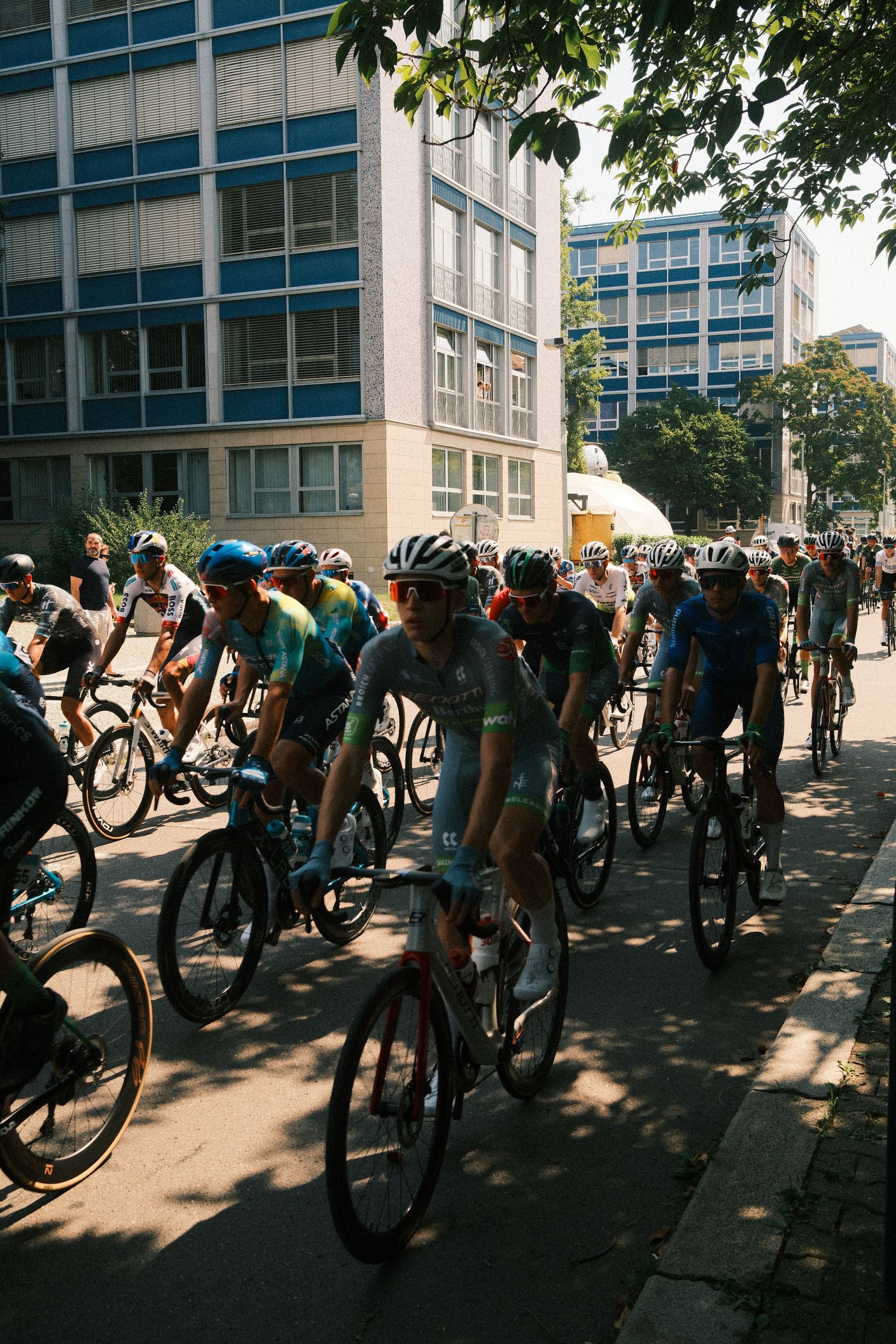 A group of cyclists riding through an urban area with buildings in the background, shaded by trees