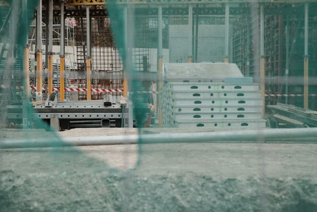 A construction site with stacked materials and scaffolding, partially obscured by green netting