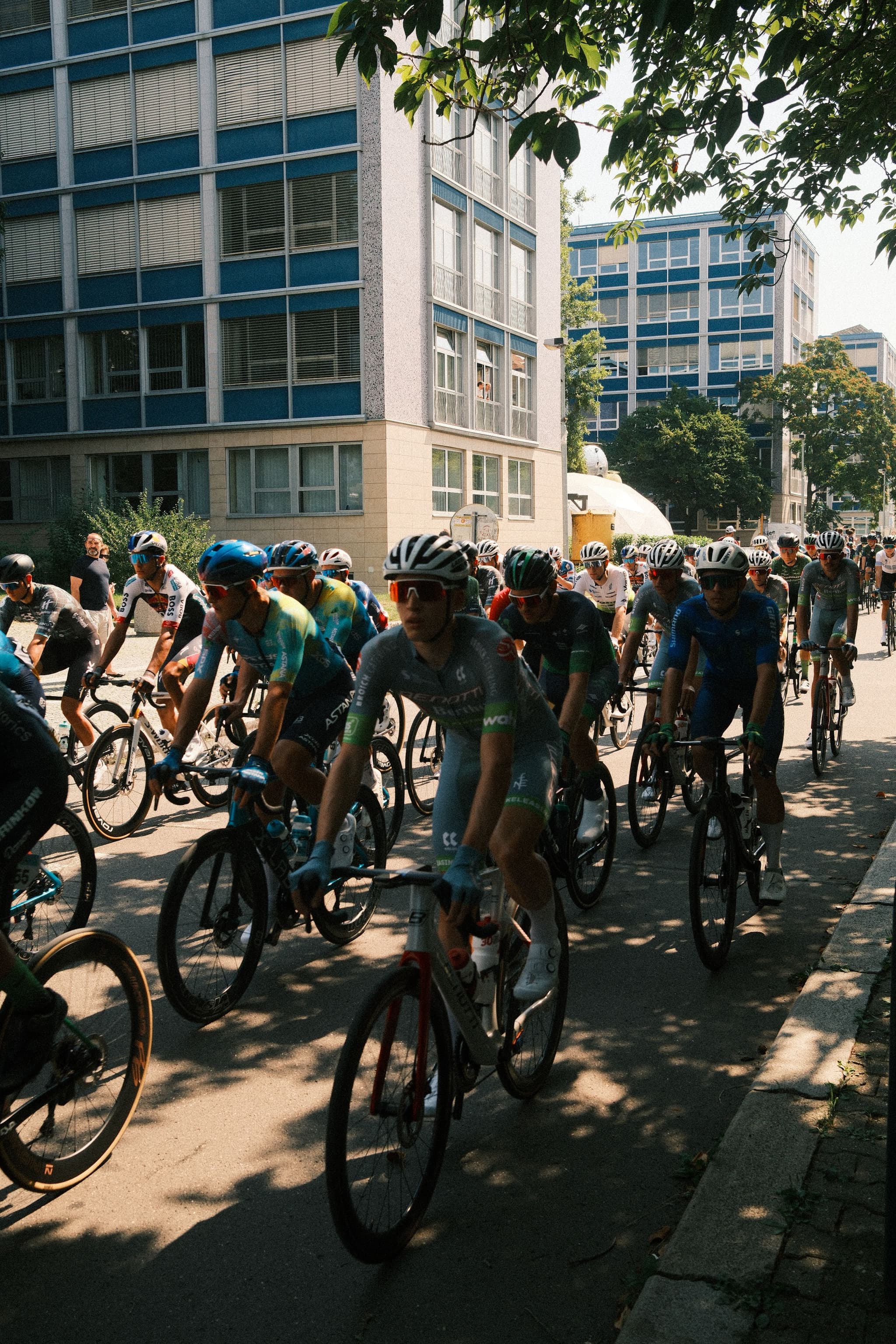 A group of cyclists riding through an urban area with buildings in the background, shaded by trees