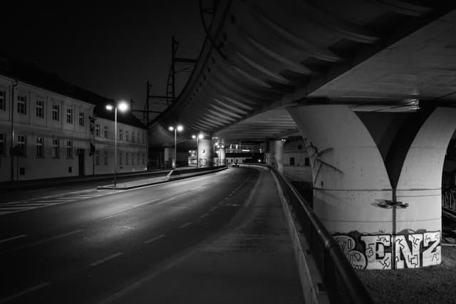 A deserted urban street at night, featuring an elevated highway with graffiti on its support pillars The scene is illuminated by streetlights, and buildings line the left side of the road
