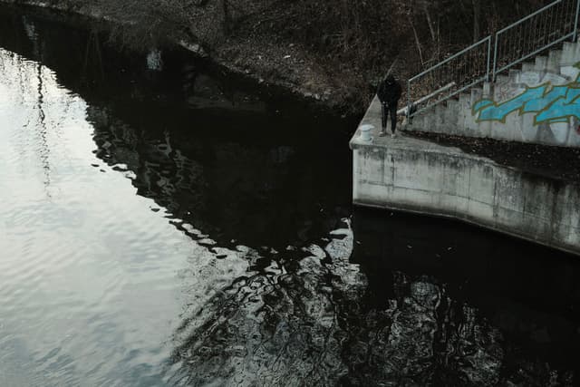 A person standing on a concrete ledge by a body of water, with graffiti on the wall and trees in the background