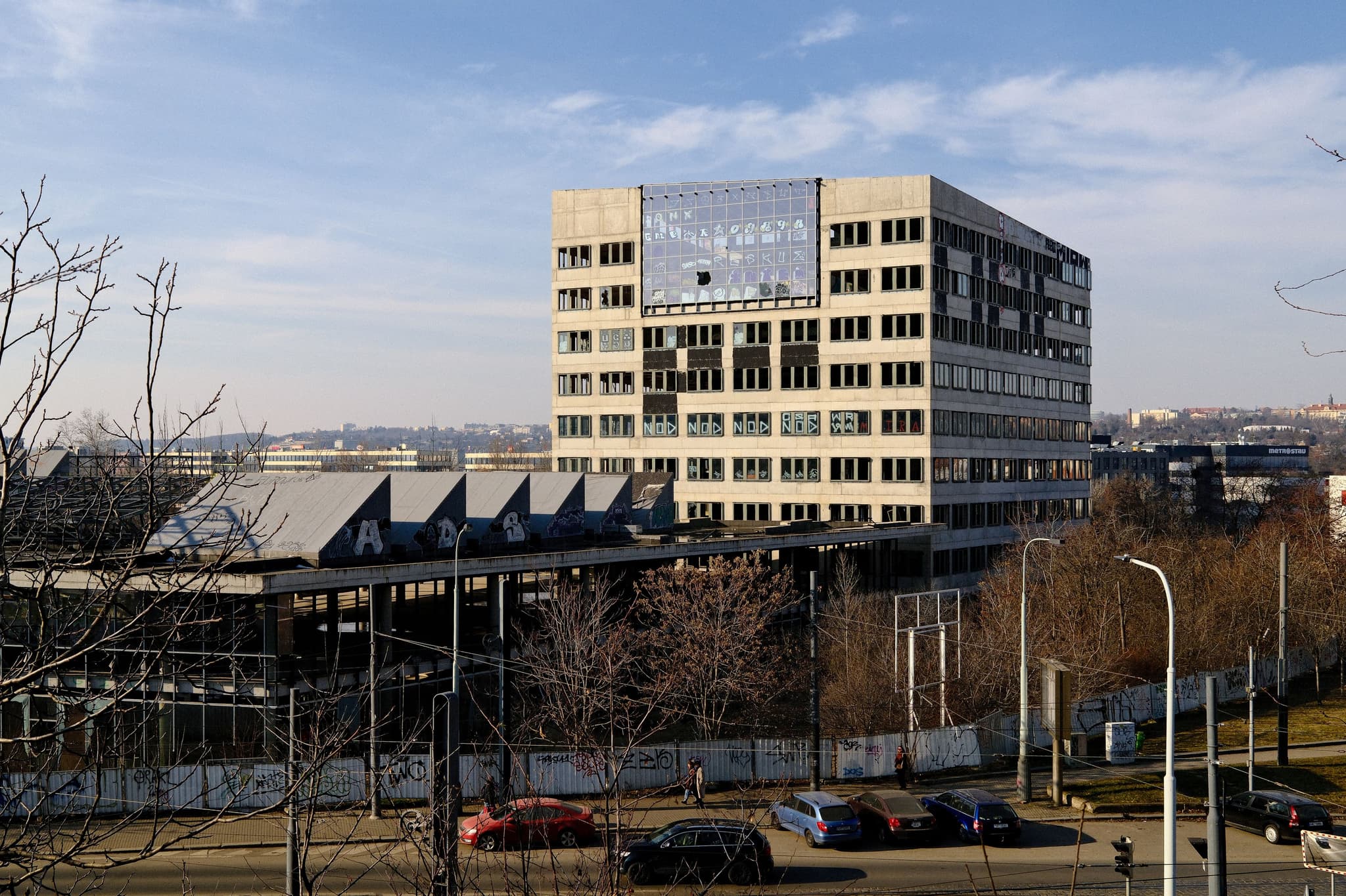A multi-story office building with a large sign on top, surrounded by trees and a fence, with a clear sky and urban landscape in the background
