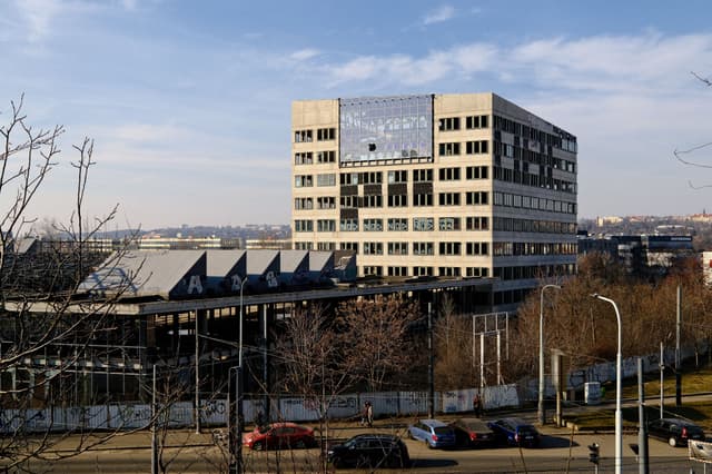 A multi-story office building with a large sign on top, surrounded by trees and a fence, with a clear sky and urban landscape in the background