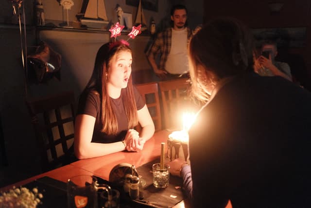 A dimly lit room with a young woman wearing star-shaped headbands, sitting at a table with a lit birthday cake in front of her Another person is leaning over the table, and a few people are in the background