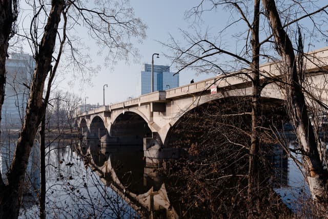 A stone bridge with multiple arches spans a calm river, surrounded by bare trees. In the background, modern buildings rise against a clear sky