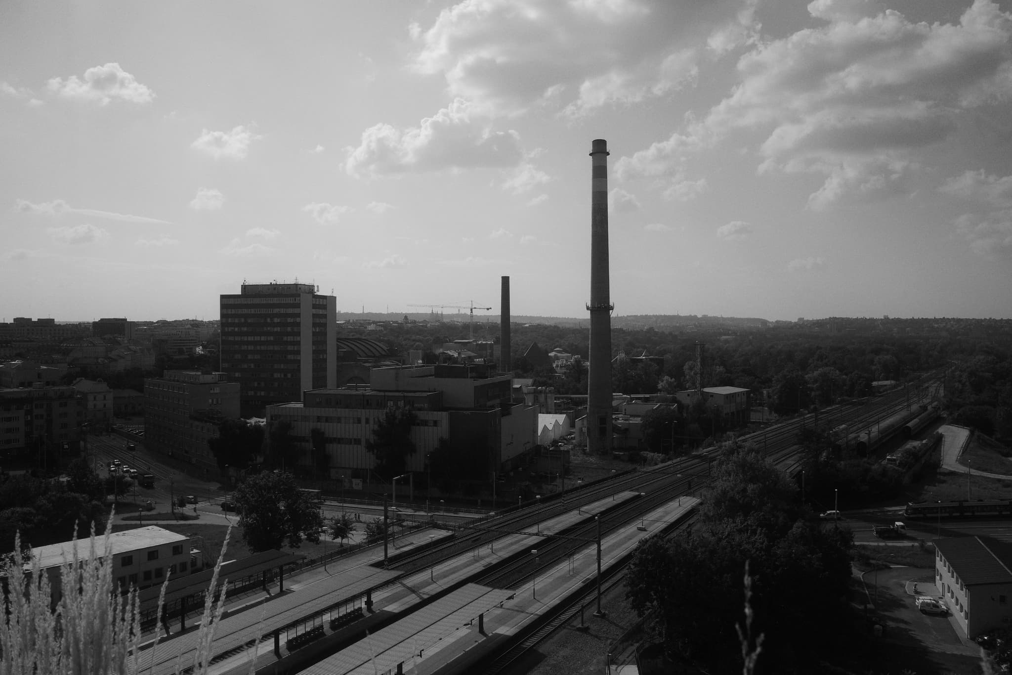 A black and white cityscape featuring tall chimneys, buildings, and a road with train tracks, under a partly cloudy sky