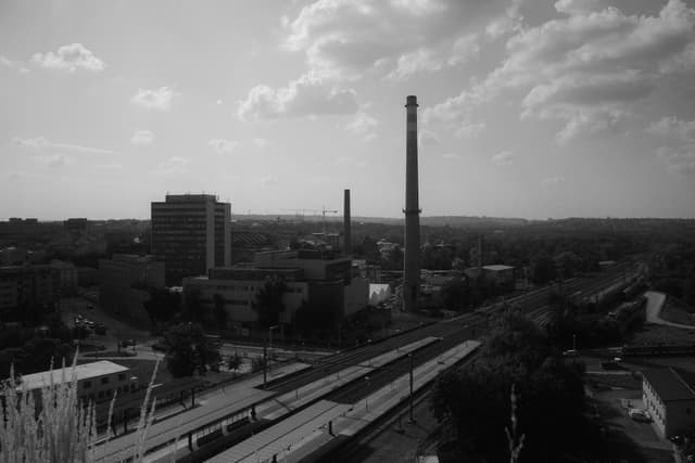 A black and white cityscape featuring tall chimneys, buildings, and a road with train tracks, under a partly cloudy sky
