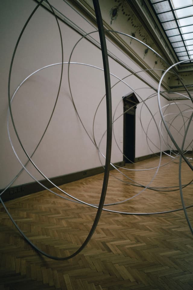 A room with large, intersecting metal rings creating an abstract installation against a wooden parquet floor and a high ceiling with skylights