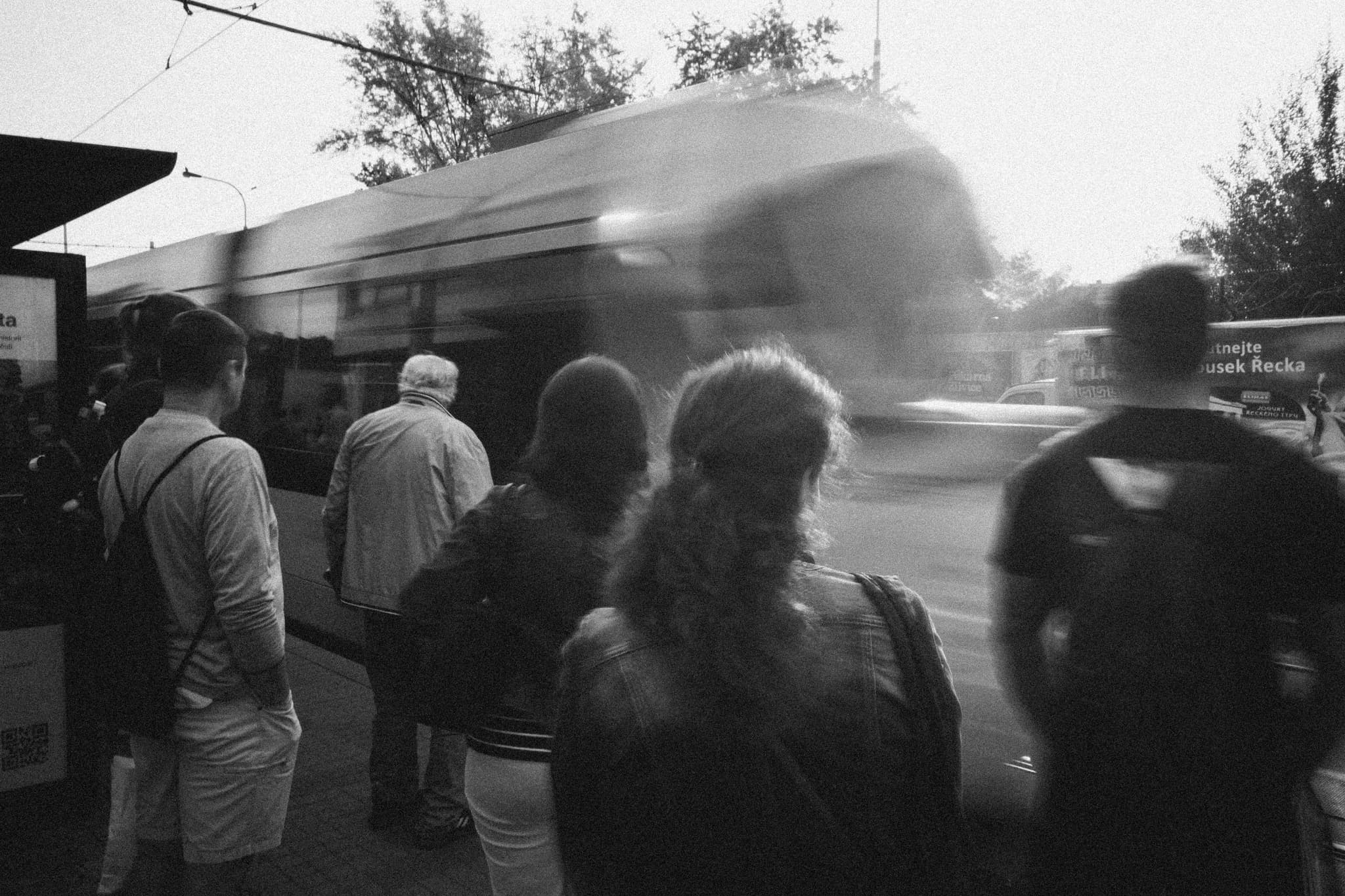 A group of people standing at a train station platform, watching a train pass by in motion blur, captured in black and white