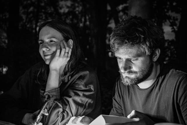 Two people sitting together in a dimly lit setting, one smiling and the other focused on reading