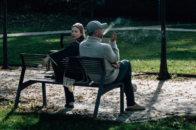 Two people sitting on a park bench, one smoking, with a grassy background and dappled sunlight